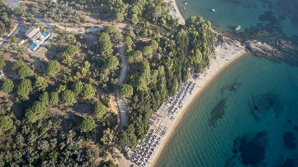 Luxurious white and blue hotel nestled on a lush Greek hillside overlooking turquoise Aegean waters, with modern minimalist architecture and elegant poolside design