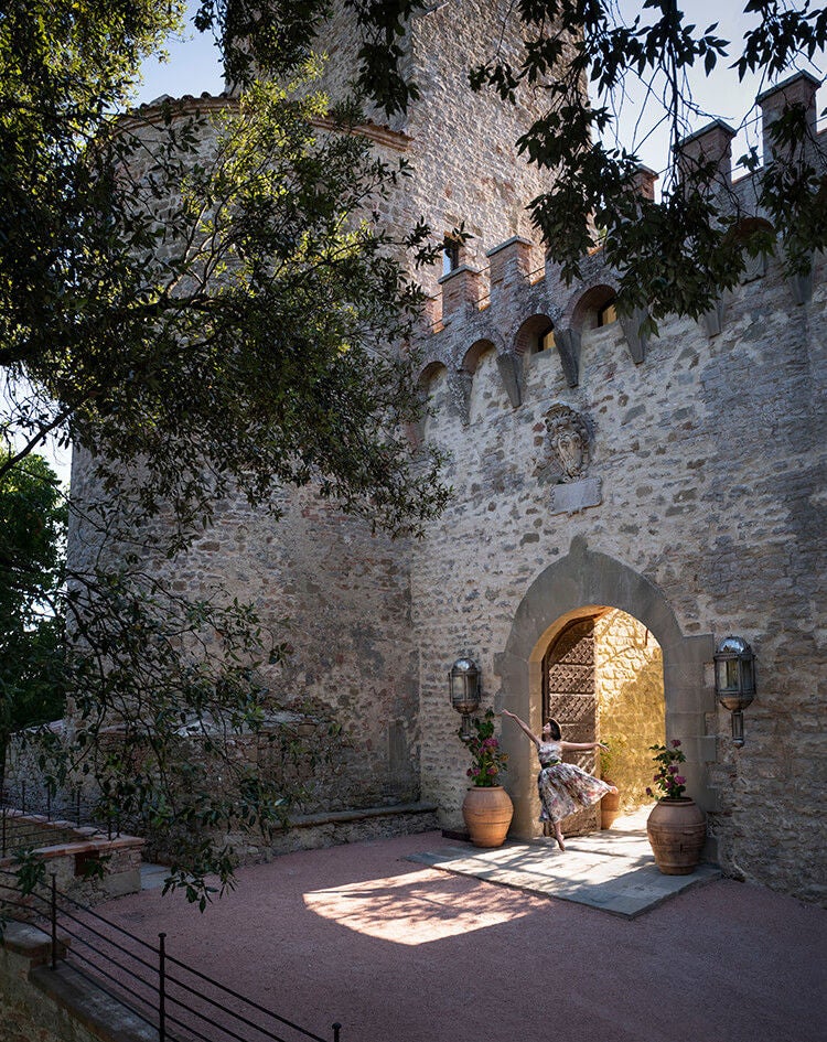 Historic Italian castle hotel with stone facade, elegant arched windows, and manicured gardens set against rolling Umbrian hills at sunset.