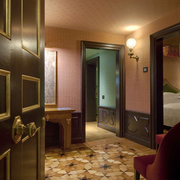 Elegant hotel suite with ornate gold-framed mirrors, crystal chandelier, plush queen bed and antique furniture in warm earth tones