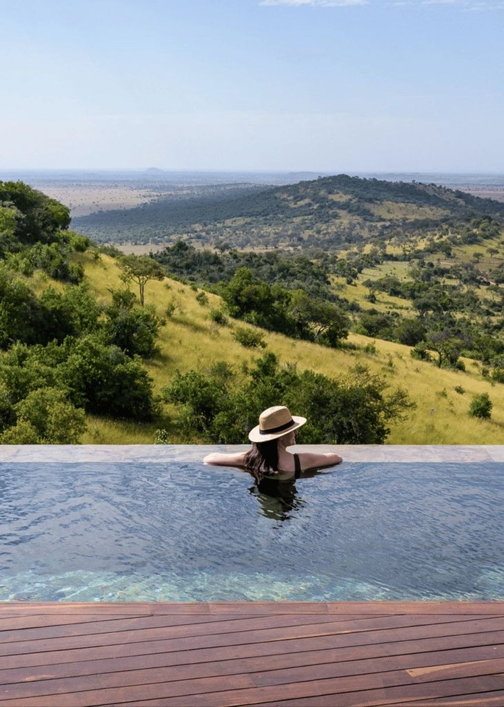 Elegant stone-built safari lodge perched on Serengeti hillside, featuring infinity pool overlooking vast savanna grasslands at sunset