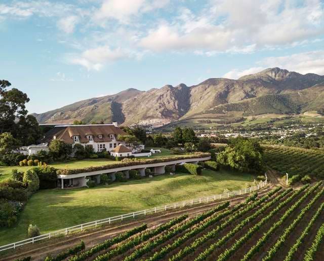 Elegant mountain-view luxury hotel nestled in South African vineyard, featuring pristine white architecture and lush green landscape with panoramic wine country backdrop