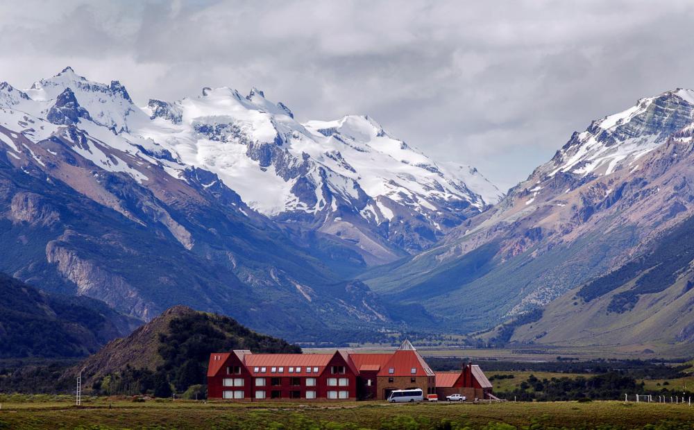 Modern mountain lodge with stone and wood facade nestled in snow-capped Andes peaks, featuring panoramic windows and rustic-luxe design