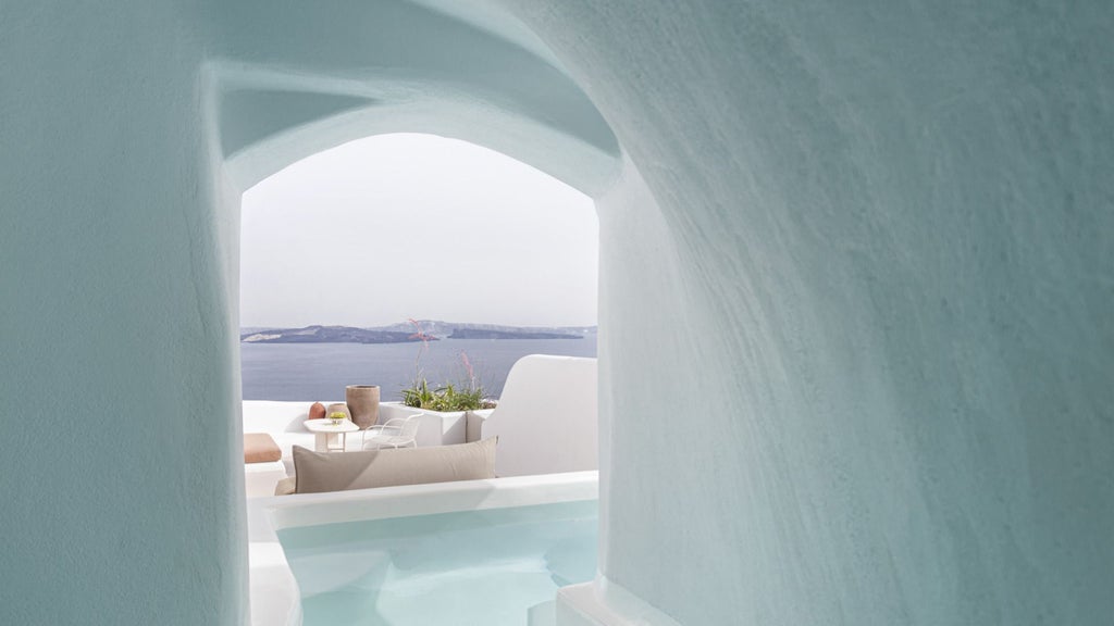 Luxurious river pool suite at Canaves Ena, featuring a minimalist white design, blue-tiled infinity pool, and panoramic view of Santorini's scenic landscape.