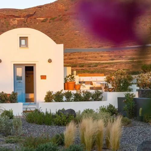 Luxurious Greek villa bedroom with white-washed stone walls, rich wooden furnishings, and panoramic Aegean Sea view through arched window at sunset