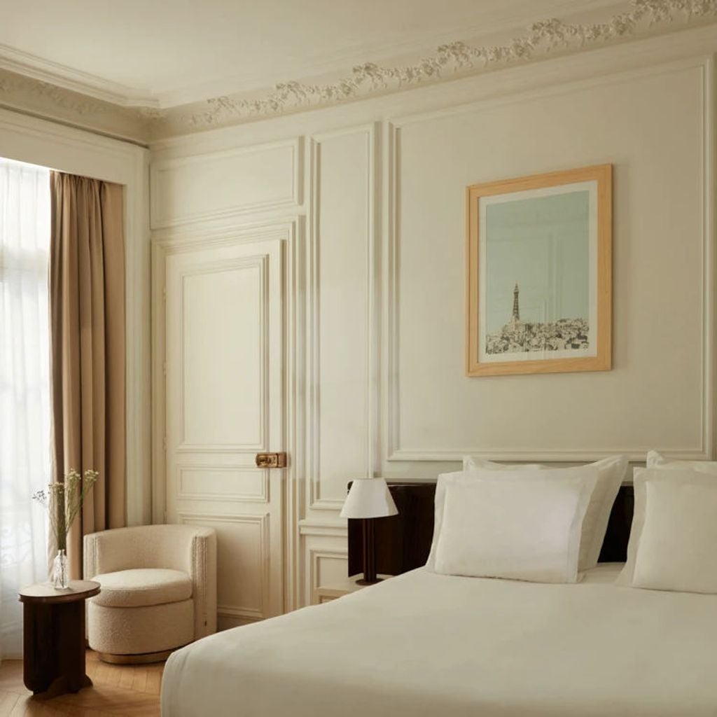 Elegant Parisian hotel room with plush king bed, cream walls, ornate moldings, chandelier, and floor-to-ceiling French windows with city views