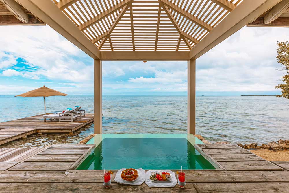 Private overwater villa with wooden deck surrounded by turquoise Caribbean waters at sunset, palm trees swaying in tropical breeze