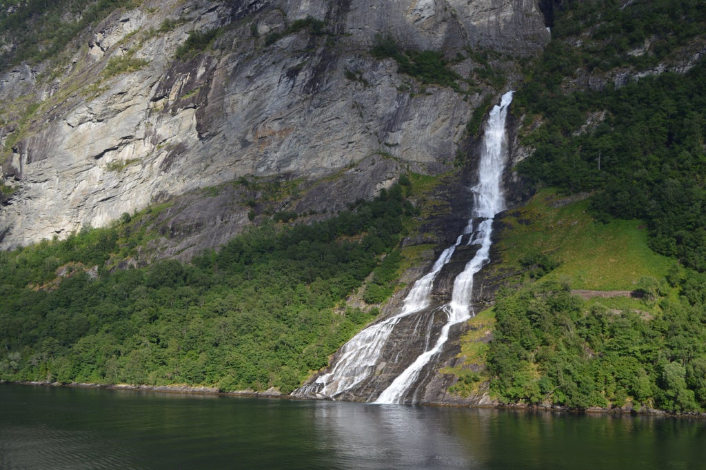 Dramatic panorama of Geirangerfjord with cascading waterfalls, lush green cliffs, and a luxury boat cruising through pristine Norwegian waters