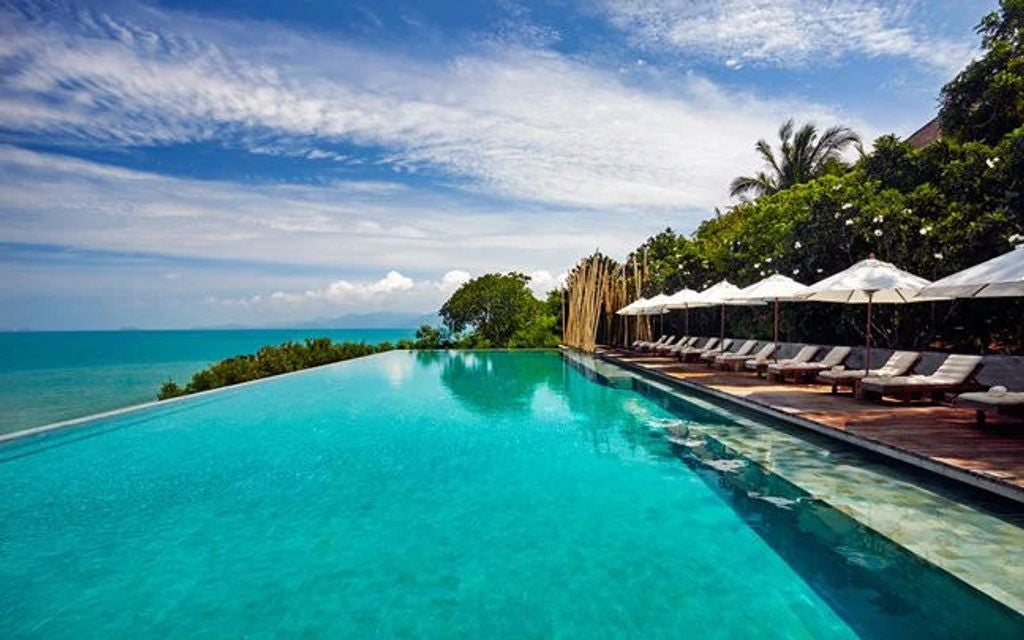 Luxurious infinity pool overlooking turquoise ocean at Six Senses Samui resort, with wooden deck and tropical palm trees at sunset