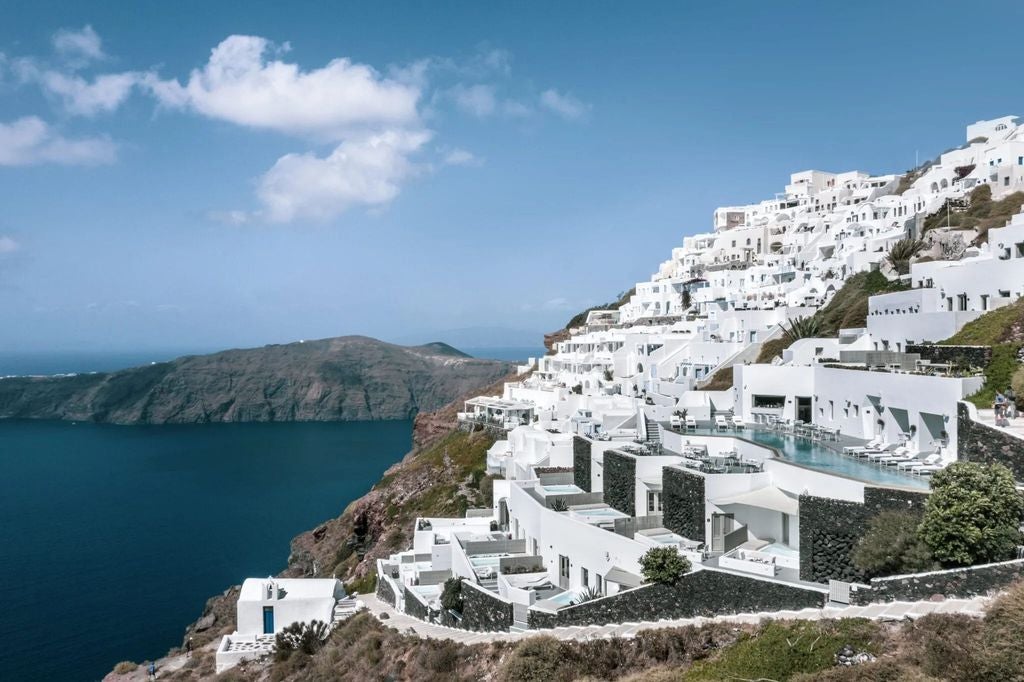 Elegant white stone hotel overlooking Santorini's caldera at sunset, with infinity pools and terraced balconies cascading down the cliff