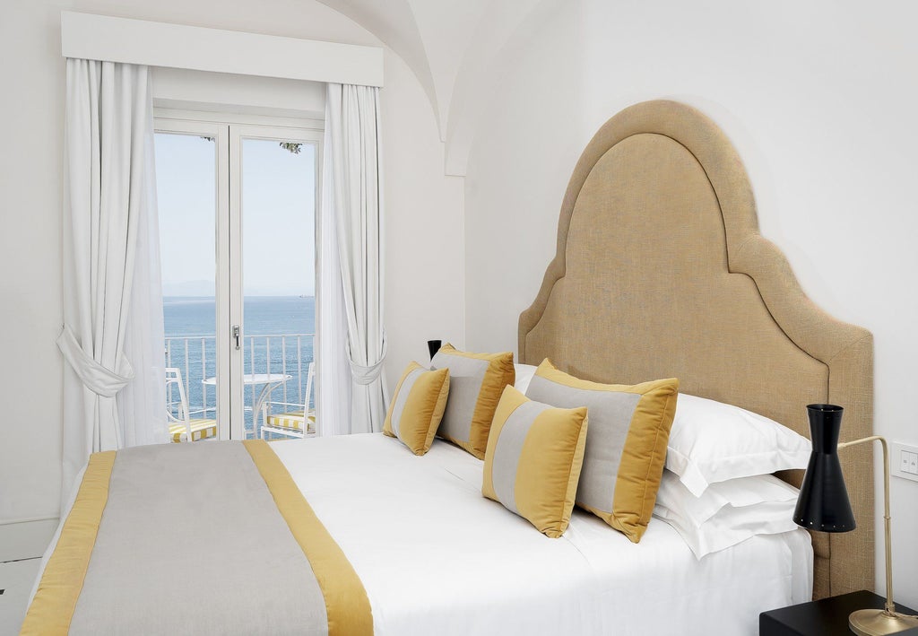 Elegant hotel room with sea-view balcony, crisp white bedding, traditional tiled floor, and blue Mediterranean vistas through French doors