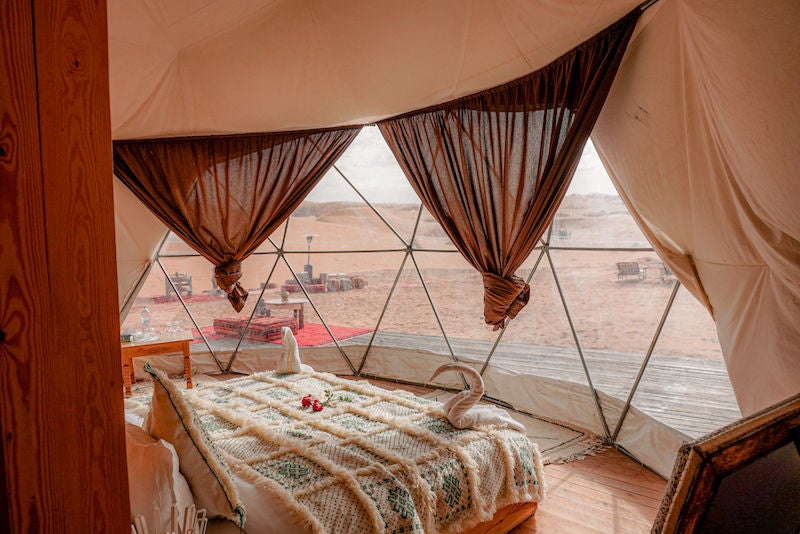 Elegant Moroccan desert camp with white canvas tents against golden sand dunes, traditional rugs and lanterns creating luxe ambiance