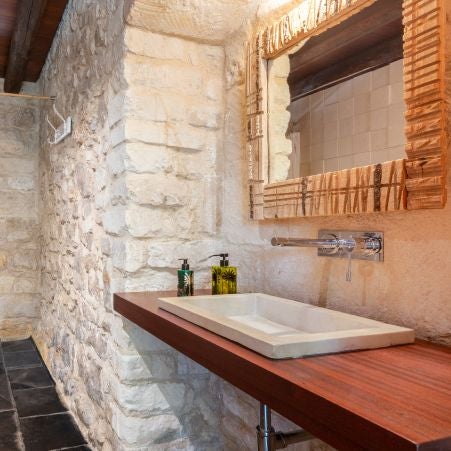 Rustic-chic Italian hotel room with stone walls, elegant wooden furnishings, soft neutral linens, and arched window overlooking scenic countryside landscape