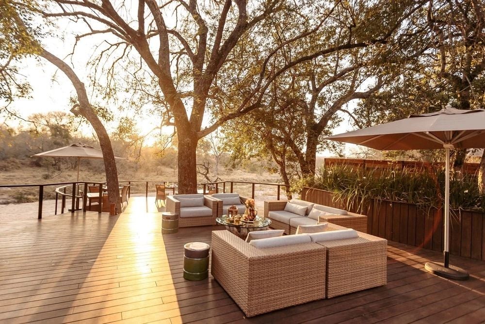 Elegant safari lodge with thatched roof and wooden deck overlooking a tranquil waterhole, surrounded by African bushveld at sunset