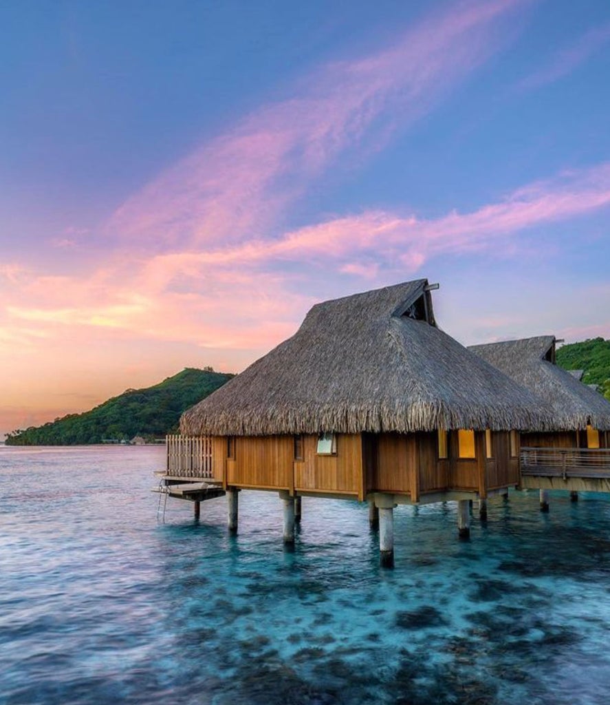 Overwater bungalows with thatched roofs stretch along turquoise lagoon at Conrad Bora Bora, backed by lush mountains and clear sky