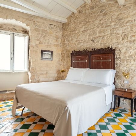 Elegant Mediterranean-style bedroom with rustic stone walls, soft white linens, and warm wooden accents at Casa Talía boutique hotel in Sicily, Italy