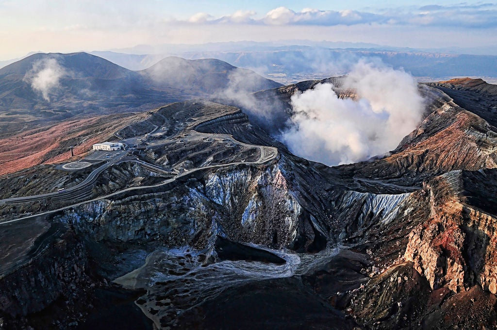 Volcanic landscape of Mt. Aso, Japan, with steaming crater, lush green slopes, and traditional architecture against a dramatic sky of soft blues and whites