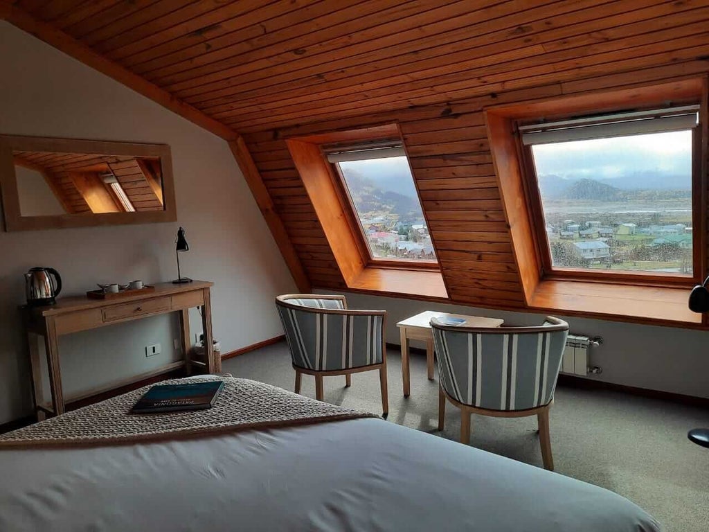 Elegant standard hotel room with warm wood accents, plush white bedding, minimalist decor, and large windows overlooking lush Argentine landscape