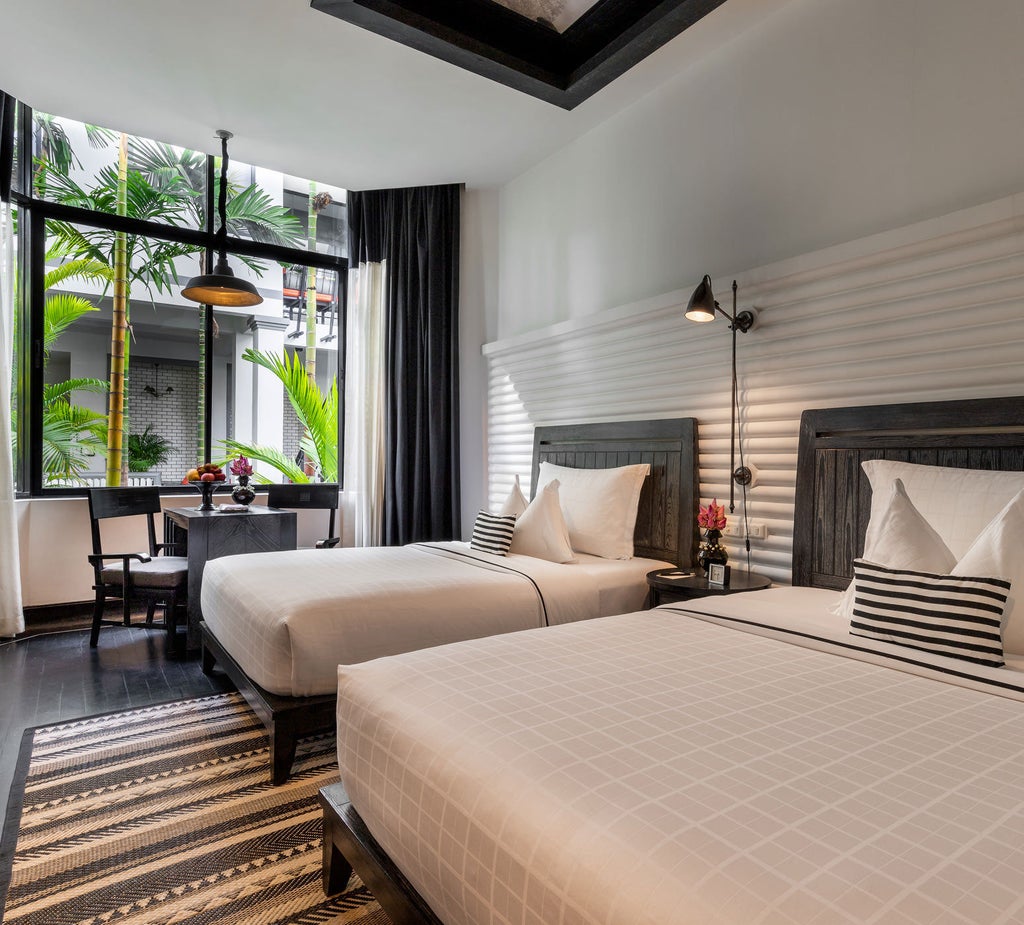 Elegant hotel room with king bed, dark wood decor, white linens, and floor-to-ceiling windows overlooking tropical Cambodian garden