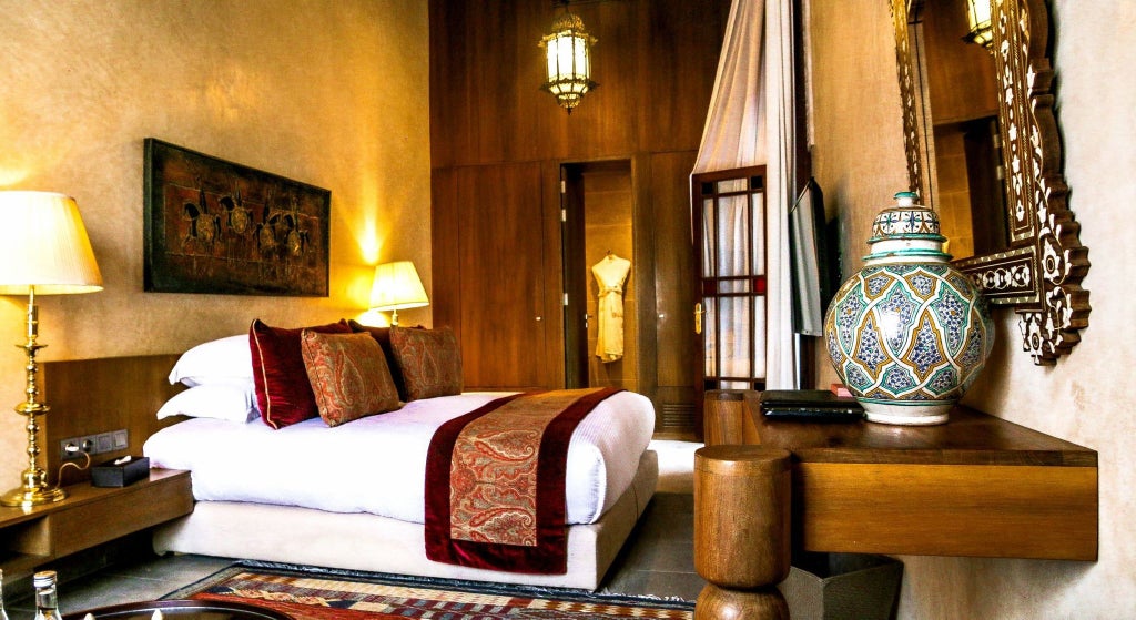 Elegant Moroccan deluxe hotel room with intricate tilework, ornate chandelier, plush white bedding, and traditional arabesque architectural details