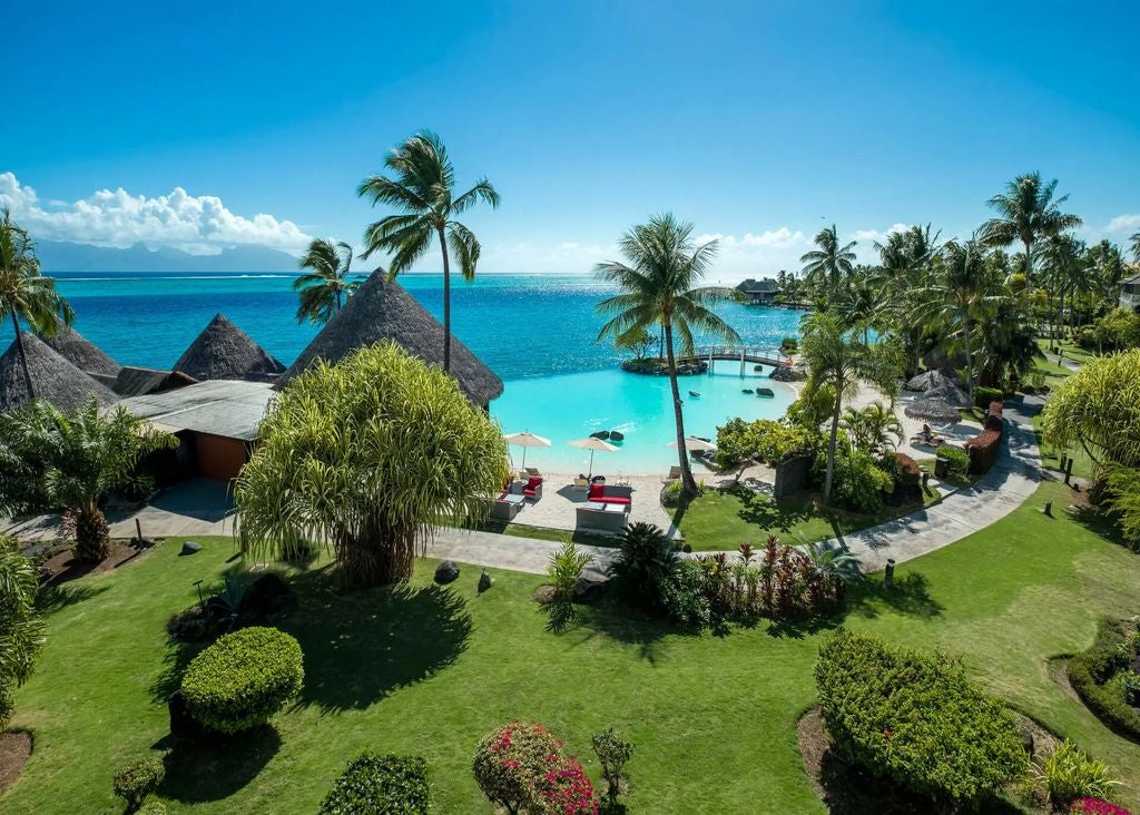 Overwater bungalows with thatched roofs stretch across turquoise lagoon, backed by lush mountains and swaying palms at sunset