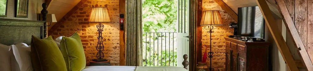 Rustic-chic hotel room with plush king bed, vintage furnishings, exposed wooden beams, and expansive windows overlooking lush countryside near Bath, UK