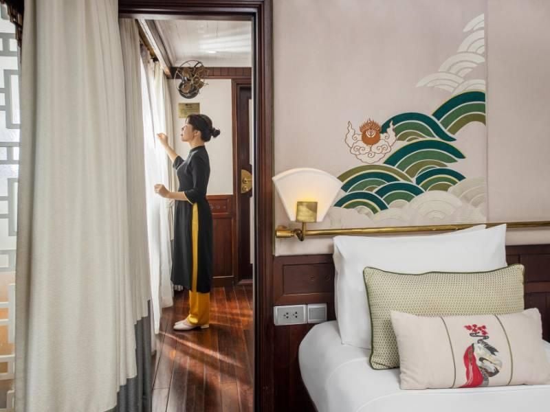 Elegant executive cabin on Bhaya Cruises, featuring rich wooden paneling, plush bedding, and panoramic windows overlooking tranquil Halong Bay waters