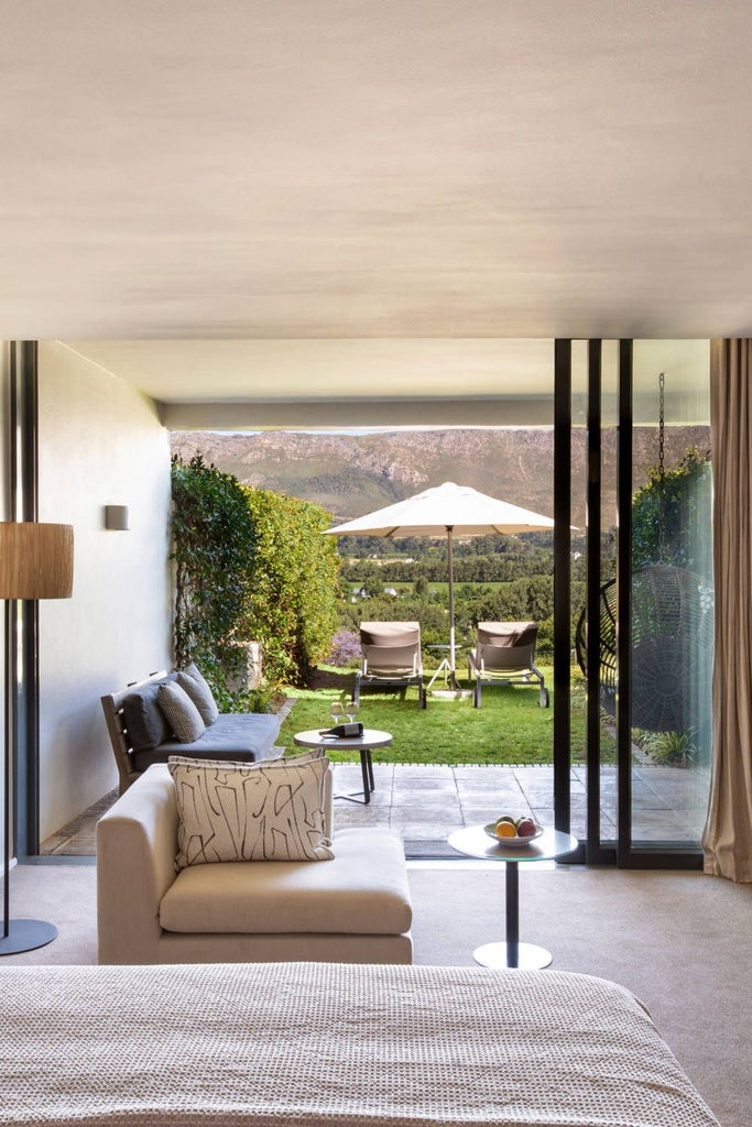 Elegant king bed with burgundy accents, plush white linens, and French doors overlooking vineyards at Mont Rochelle luxury room in South African wine country