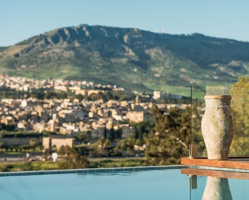 Elegant Hotel Sahrai's exterior in Morocco features modern white architecture, infinity pool overlooking city, and mountain views at sunset