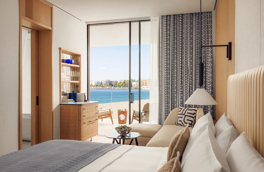 Luxurious minimalist hotel room with white linens, sleek modern furniture, expansive floor-to-ceiling windows overlooking scenic Greek coastal landscape