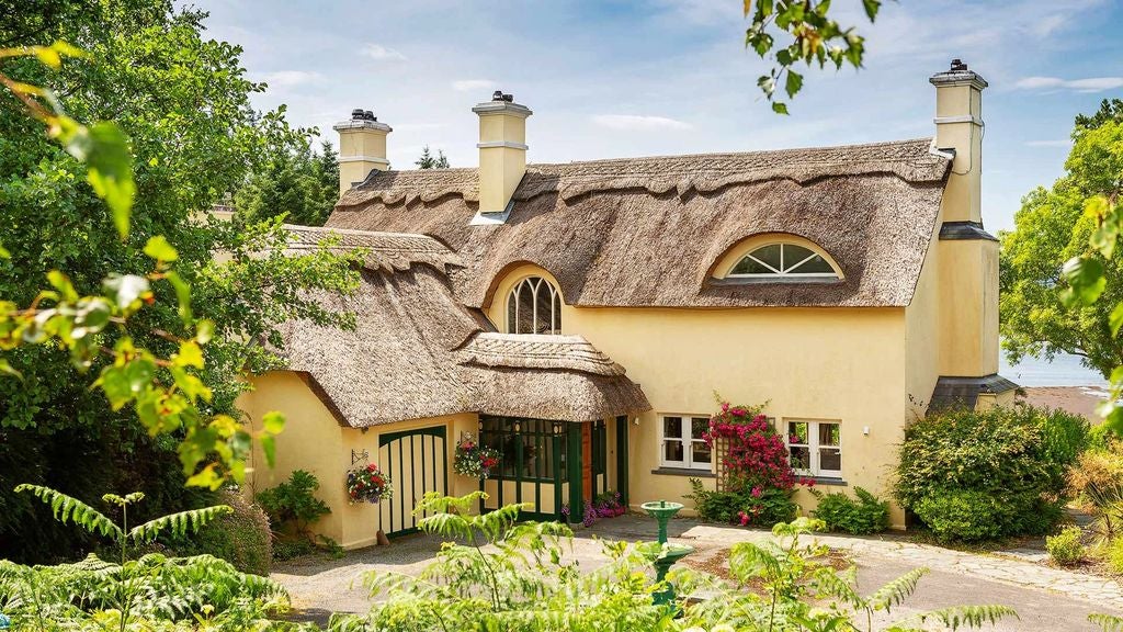 Spacious cottage bedroom with elegant four-poster bed, rich wooden beams, plush armchairs and classic Irish countryside views through windows