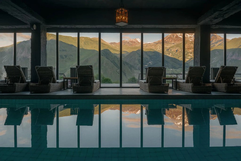 Mountain-view luxury hotel room with modern minimalist design, wooden floors, large windows overlooking dramatic Caucasus mountain landscape at sunset