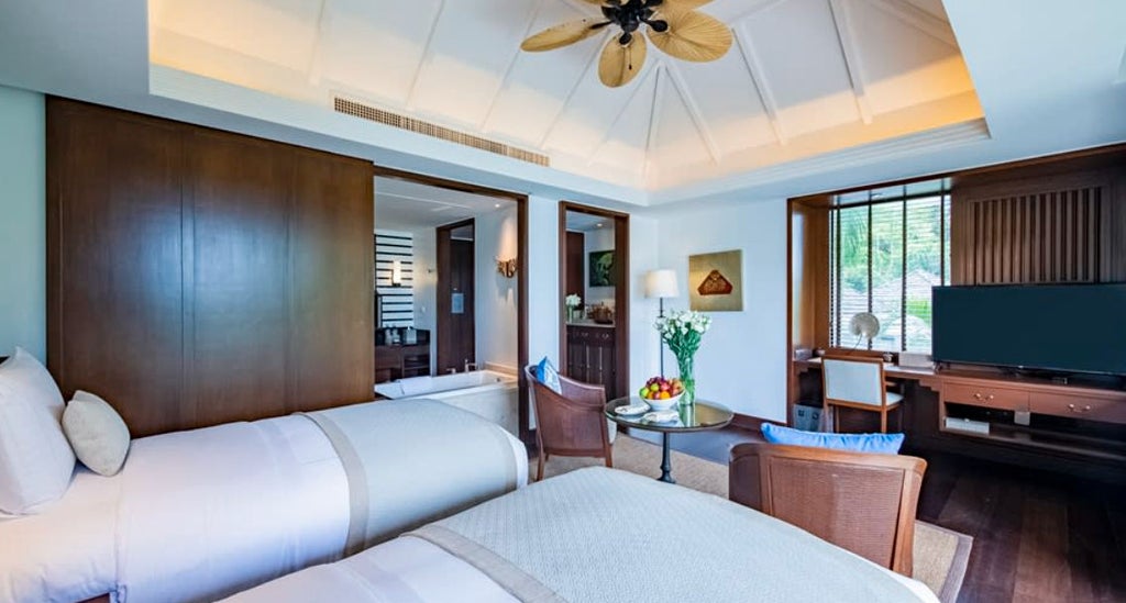 Spacious luxury hotel room with king bed, floor-to-ceiling windows overlooking tropical gardens, hardwood floors, and modern Thai decor