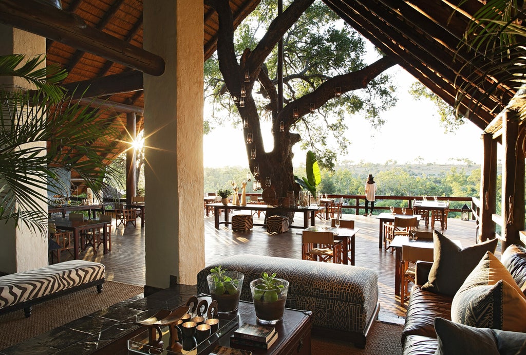 Elegant safari lodge with thatched roof overlooks private deck and infinity pool, surrounded by lush African bush at sunset