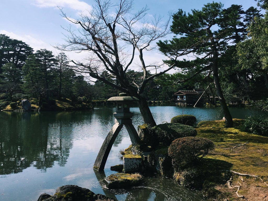 Traditional Japanese garden at Kenroku-en featuring stone lanterns, manicured trees, and a serene pond with autumn maple leaves.