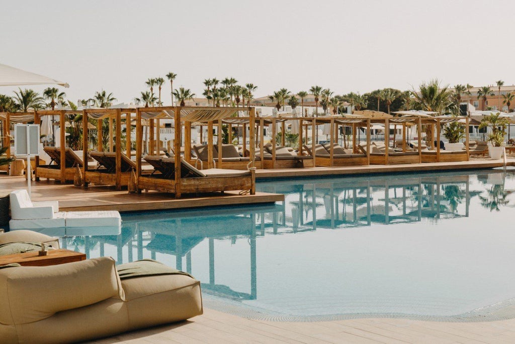 Luxurious Mediterranean beachfront resort with white buildings, azure pool, and palm trees overlooking turquoise waters of Menorca's scenic coastline