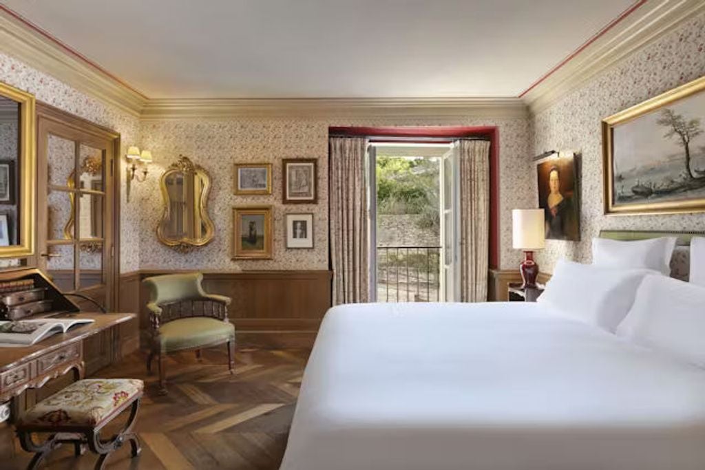 Elegant French country hotel room with exposed stone walls, ornate headboard, crystal chandelier, and panoramic village views