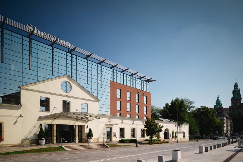 Sheraton Grand Krakow's elegant glass facade reflects blue sky, overlooking Vistula River with stone promenade and outdoor terrace