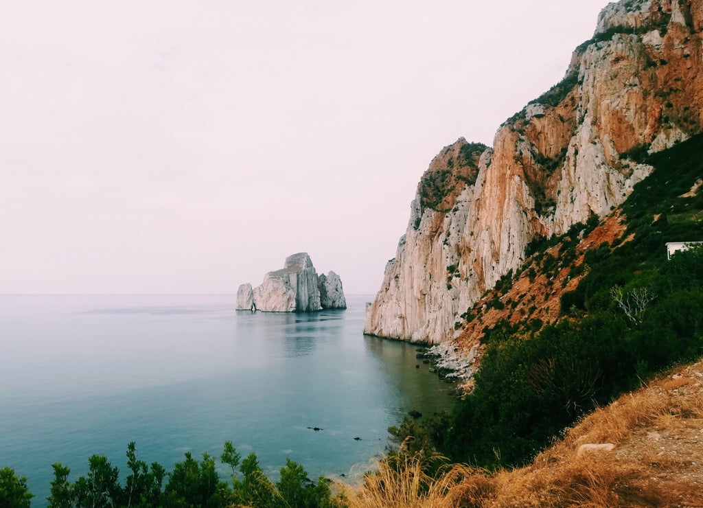 Crystal-clear turquoise Mediterranean waters meet rugged cliffs along Sardinia's pristine coastline, with luxury yachts anchored in a secluded bay