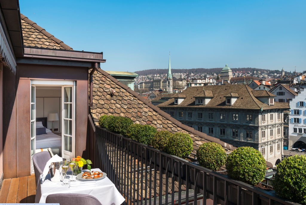 Elegant riverside suite with floor-to-ceiling windows, plush king bed, modern chandelier and panoramic views of Zurich's waterfront