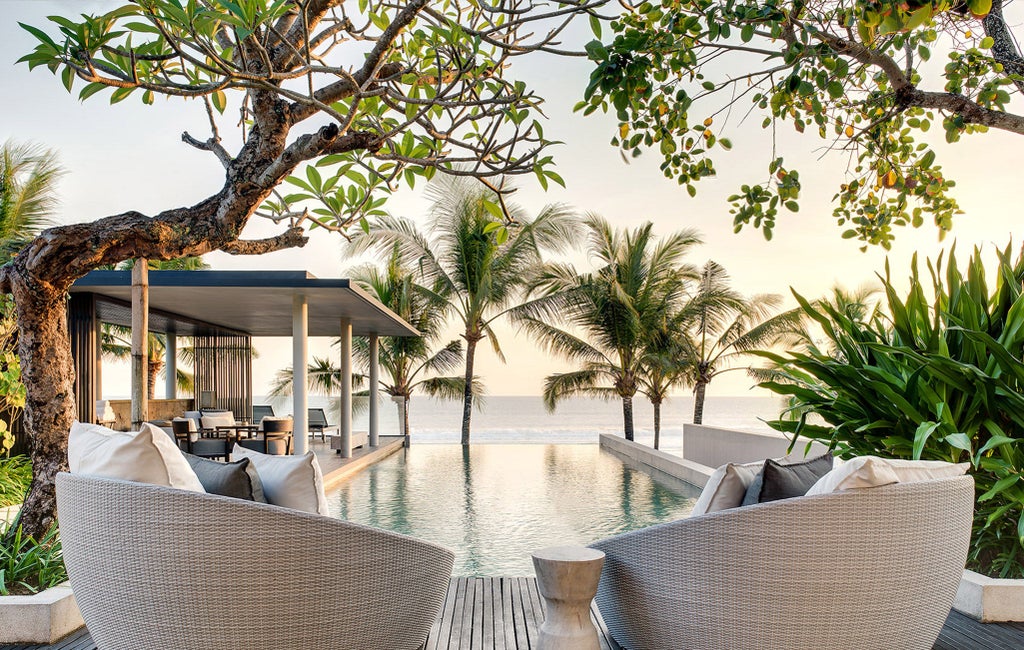 Luxury beachfront infinity pool surrounded by minimalist stone architecture merging with dramatic ocean views at sunset in Bali