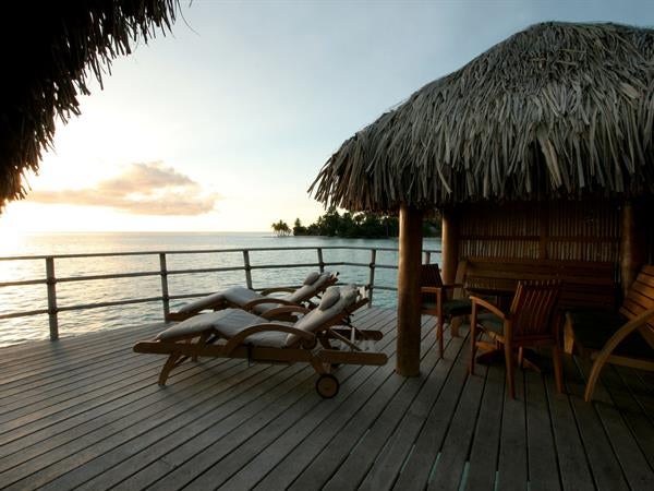 Luxurious overwater villa with private deck extending over crystal-clear lagoon, featuring thatched roof and stunning sunset views