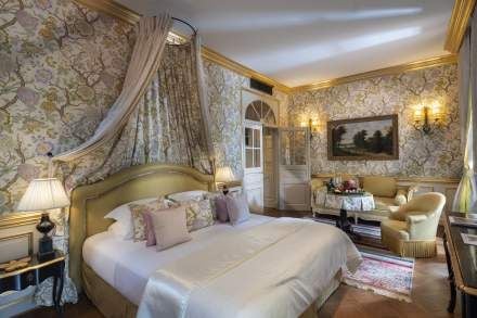 Elegant Provençal-style junior suite with soft blue-grey decor, plush white bedding, antique furnishings, and french windows overlooking lavender gardens