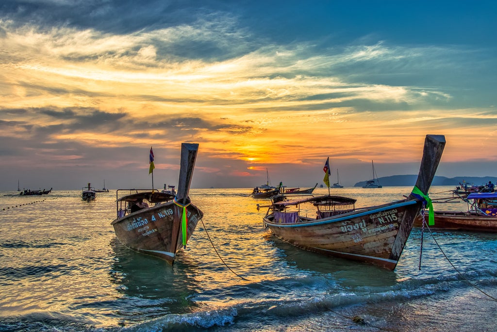 Thai fisherman steering traditional wooden longtail boat through calm river, with lush mangroves and rustic village houses on stilts along shore