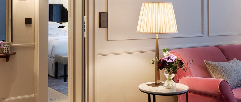 Elegant Swedish hotel suite with plush king bed, warm wood tones, minimalist Nordic design, soft neutral color palette, and large window overlooking cityscape