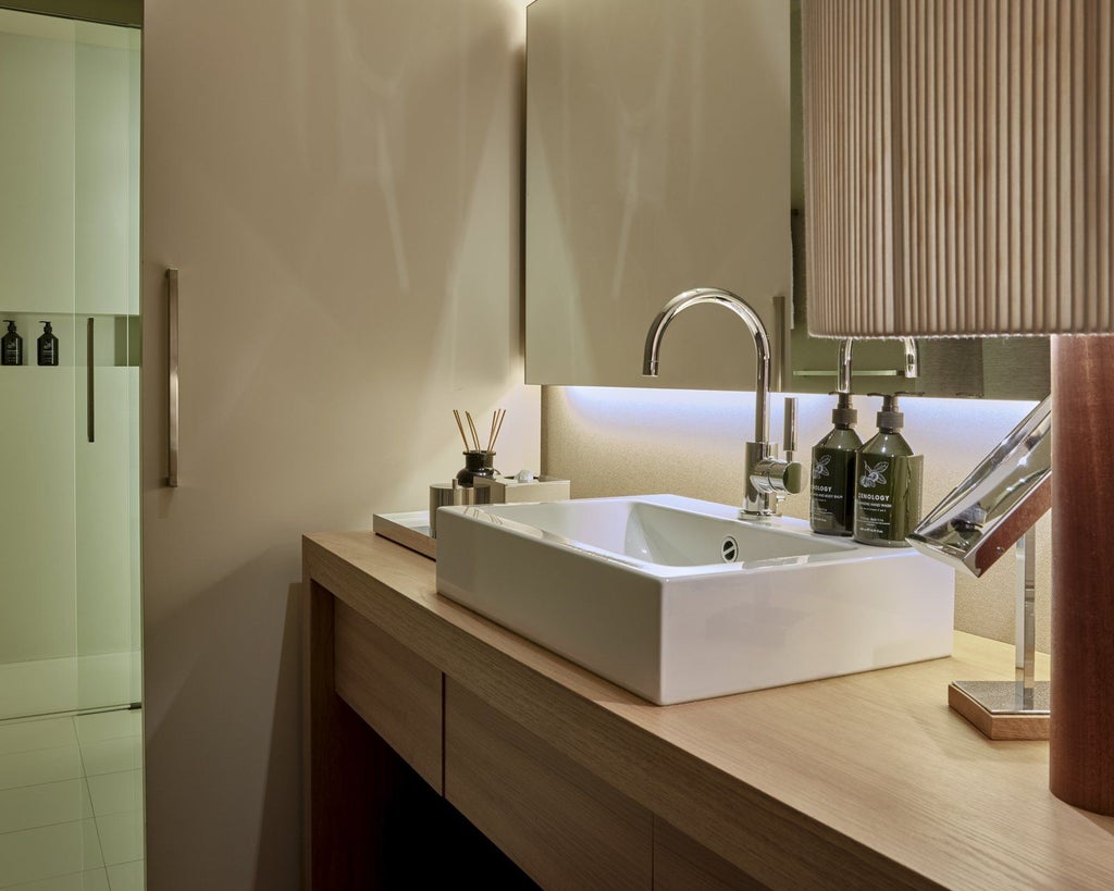 Modern luxury hotel room with minimalist design, wooden flooring, white linens, and panoramic window overlooking urban Barcelona skyline at sunset