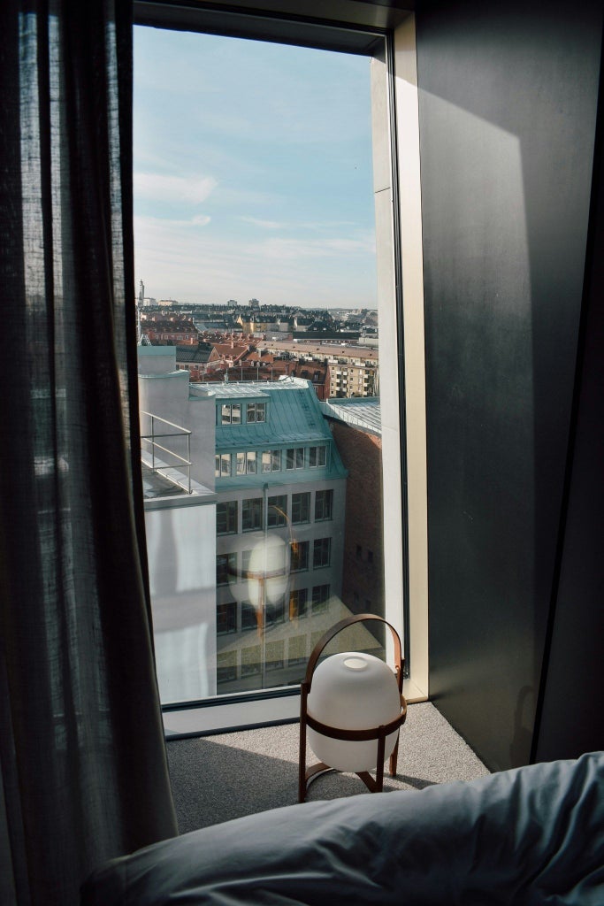 Modern minimalist Scandinavian hotel studio with floor-to-ceiling windows, sleek gray furnishings, and natural light overlooking urban Stockholm landscape