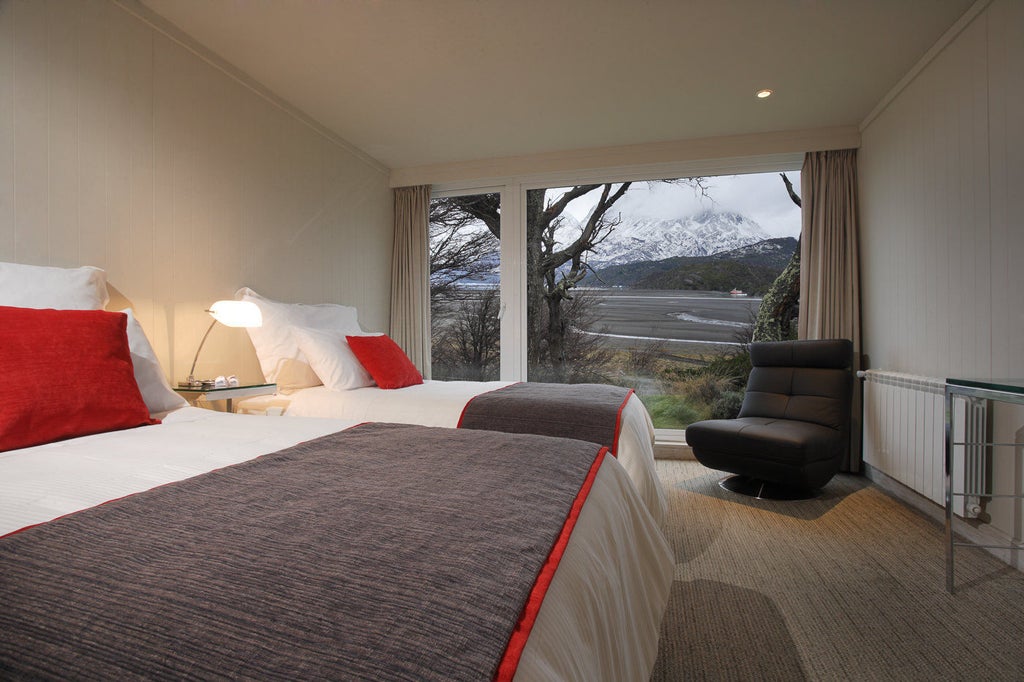 Modern mountain lodge surrounded by snow-capped peaks, featuring wood and glass facade, overlooking grey glacial lake in Torres del Paine