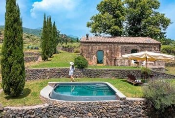 Elegant Sicilian estate suite with rustic stone walls, minimalist decor, and large windows overlooking lush volcanic landscape at Monaci delle Terre Nere