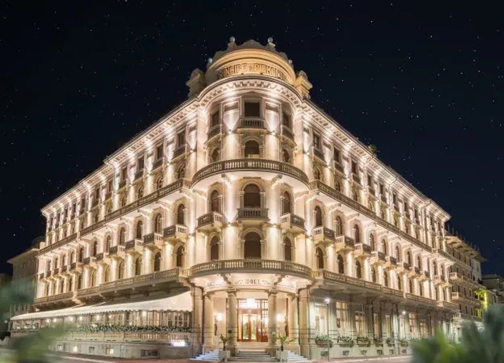 Opulent Italian hotel exterior with marble façade, ornate balconies and classic Mediterranean architecture set against clear blue sky