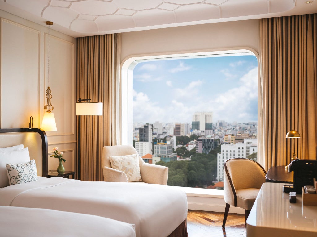 Modern twin hotel room with floor-to-ceiling windows offering city views, featuring plush beds, elegant furnishings, and warm lighting